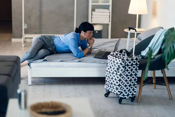Woman lying on bed working remotely using laptop computer indoors