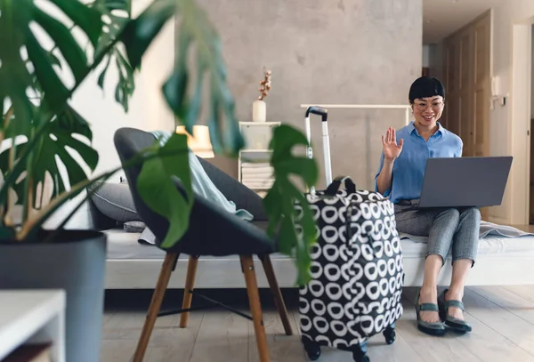 Smiling woman sitting on bed using laptop waving near suitcase indoors