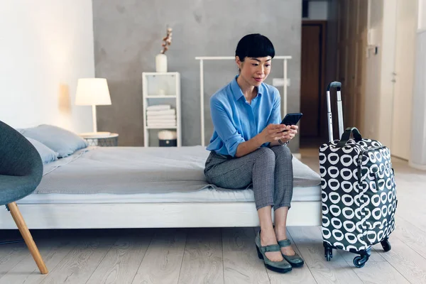 Smiling woman sitting on bed checking phone near suitcase