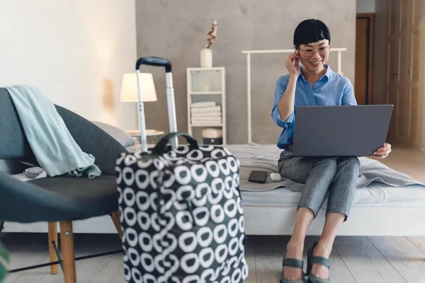 Smiling woman sitting on bed using laptop while packing suitcase nearby