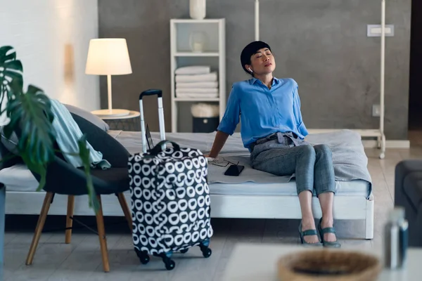 Tired woman resting on bed after arriving with patterned suitcase