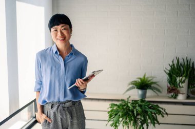 Smiling woman holding tablet standing near indoor green plants