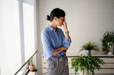 Woman standing indoors touching face looking stressed near green plants