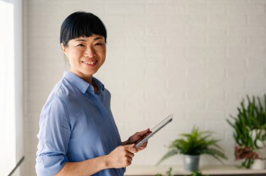 Smiling woman holding tablet standing near green indoor plants