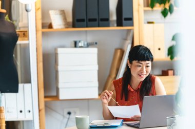 Smiling woman working at laptop holding paper in creative office