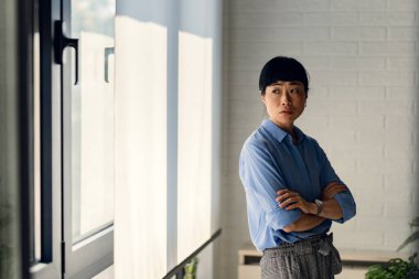 Confident woman standing near window crossing arms indoors