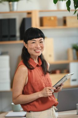 Smiling woman holding digital tablet standing near office shelves