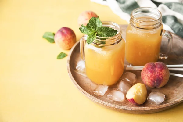 Cold summer peach drink with ice and mint in mason jars on a tray with fresh peaches on yellow background. Copy space.