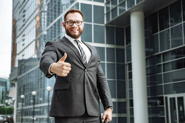 Портрет счастливого бизнесмена, показывающего большой палец на фоне бизнес-центра — стоковое фото
