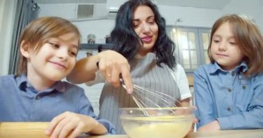 Mutlu aile mutfakta krep ya da kurabiye için hamur hazırlıyor. Genç anne çırpılmış yumurta çırparken tatlı çocuklar gülüyor ve zıplıyor evde birlikte eğleniyorlar..