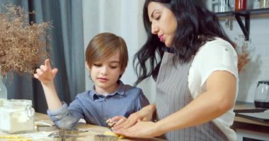 Genç bir anne oğluyla ev yapımı kurabiyeler hazırlıyor hamuru yoğuruyor ve mutfaktaki kurabiye kesicilerle figürleri kesiyor..