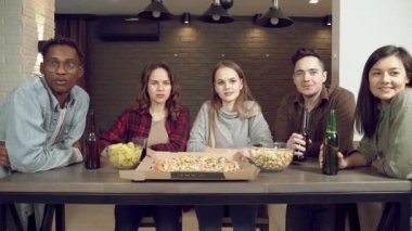 Bir grup mutlu çoklu etnik arkadaş pizza yiyip, bira içerken birlikte televizyon izliyorlar..