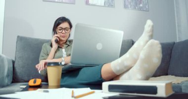 Genç bir kadın, iş arkadaşları ya da müşterileriyle akıllı telefondan konuşuyor, bir projeyi tartışıyor ve evdeki bir kanepede oturup dizüstü bilgisayarda çalışarak notlar alıyor.