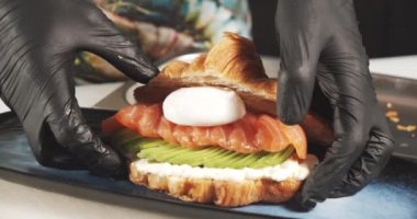 Yakından aşçının elleri eldivenli lezzetli bir kruvasan, peynir, avokado, somon filetosu ve yumurtalı Benedict pişiriyor. Lezzetli ve sağlıklı kahvaltı..