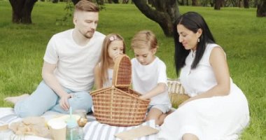 Yeşil bahçedeki piknik sırasında çimlerin üzerinde dinlenen mutlu bir aile. Küçük çocuk sepetten lezzetli çörekleri alıp masaya koyuyor..