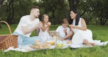Mutlu ve çocuklu bir aile yeşil bahçede piknik yaparken çimenlerde dinleniyor ve lezzetli çörekler yiyor.