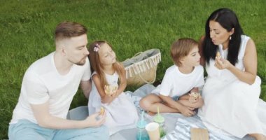 Mutlu ve çocuklu bir aile yeşil bahçede piknik yaparken çimenlerde dinleniyor ve lezzetli çörekler yiyor..