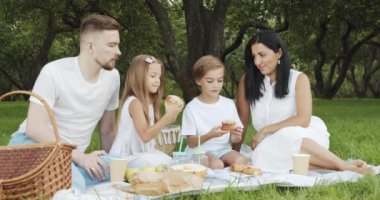 Mutlu ve çocuklu bir aile yeşil bahçede piknik yaparken çimenlerde dinleniyor ve lezzetli çörekler yiyor.