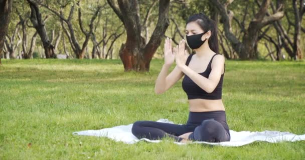 晴れた日に公園で黒い保護マスクを身に着けている間 若い女性は蓮の位置でヨガ瞑想を練習します 流行で健康的なライフスタイルのコンセプト — ストック動画