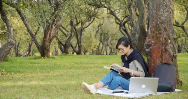 公園の芝生に座ってセミナーの準備中に本を読んで眼鏡をかけた若い女性学生 — ストック動画