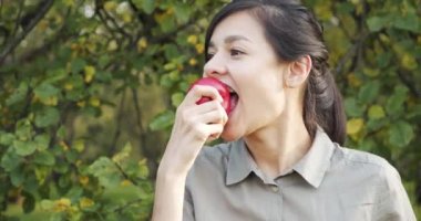 Elma yiyen ve yeşil bahçedeki kameraya bakan çekici bir kadının portresi. Bahçıvanlık ve tarım kavramı. Organik yiyecekler.