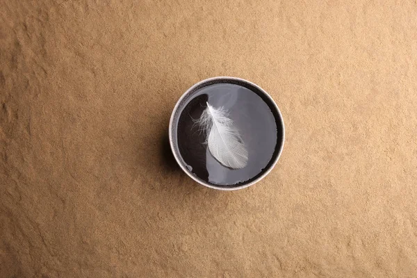 White feather on water surface in bowl on sand background. Ceramic  bowl with clean drink water and feather outdoors, top view.