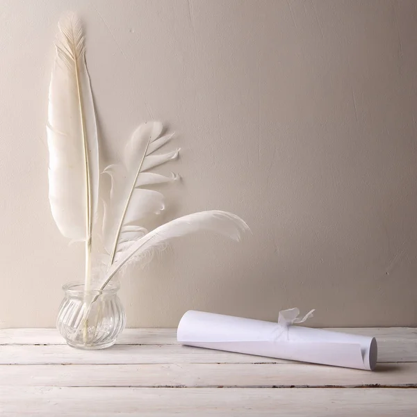 Paper rollers and white quill feathers in glass bottle on white wood table. Feathers and paper rollers against white wall with empty place.
