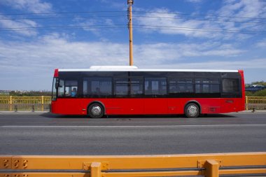 Şehir otobüsü köprüden geçiyor. Turuncu bariyer veya korkuluk lara karşı yolda hareket eden kırmızı otobüs.