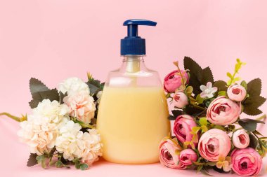 Sarı sıvı sabun şişesi ve pembe arka planda güzel narin çiçekler. Temiz eller kavramı. Şampuan, Sıvı Sabun, Aromatik Banyo Tuzu ve Diğer Tuvaletler.