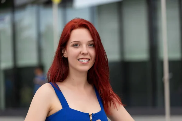 Pretty young woman with a wonderful sense of humor, red hair and a blue dress on a city street laughing at the camera