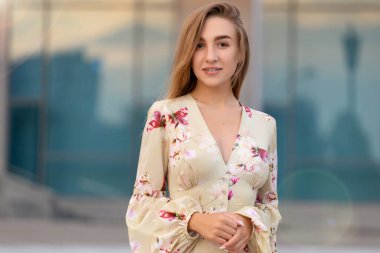 Mavi bir iş merkezinin arka planında bej bir elbise giymiş güzel bir genç kızın portresi.