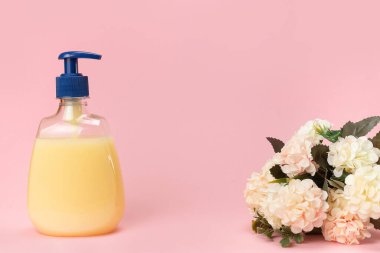 Sarı sıvı sabun şişesi ve pembe arka planda güzel narin çiçekler. Temiz eller kavramı. Şampuan, Sıvı Sabun, Aromatik Banyo Tuzu ve Diğer Tuvaletler.