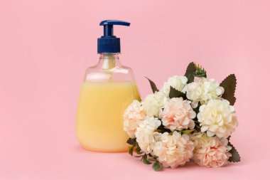 Sarı sıvı sabun şişesi ve pembe arka planda güzel narin çiçekler. Temiz eller kavramı. Şampuan, Sıvı Sabun, Aromatik Banyo Tuzu ve Diğer Tuvaletler.