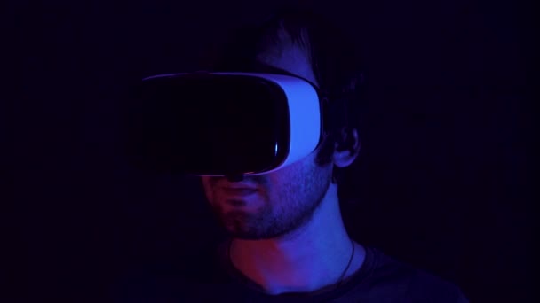 Young man in VR headset looks around and wonders how amazing. Virtual reality helmet on black technology background. Modern fluorescent light — Stock Video