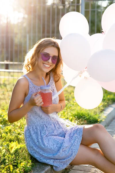 Chica feliz sosteniendo globos de aire y bebidas frías mientras está sentado en el suelo — Foto de Stock