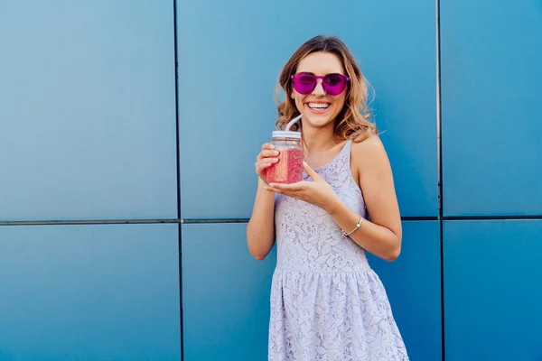 Mujer sonriente en gafas de sol bebiendo bebida de frutas, de pie contra la pared azul — Foto de Stock