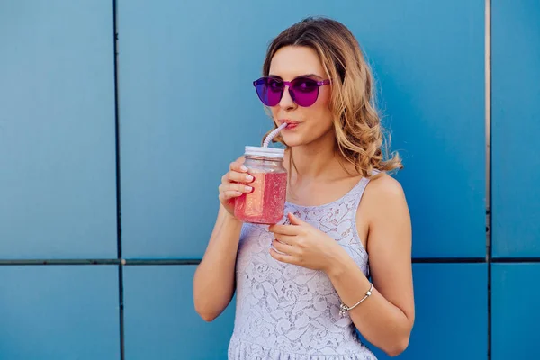 Linda chica en gafas de sol bebiendo batido fresco a través de una paja, al aire libre . — Foto de Stock