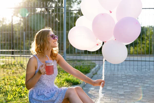 Boční pohled na šťastnou dívku držící vzduchové balónky a studený nápoj při sezení na trávě — Stock fotografie