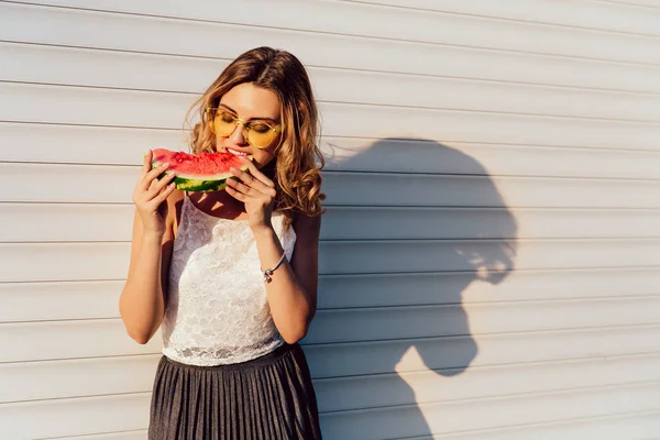 Increíble chica comiendo una sandía, de pie cerca de la pared, al aire libre . — Foto de Stock