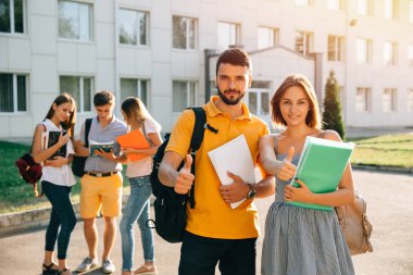 Not defteri ve sırt çantaları ile mutlu iki genç öğrenci gülümseyerek ve başparmak gösteren