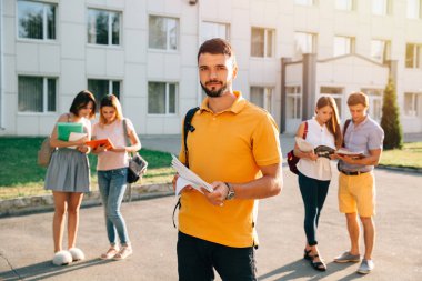 Üniversite ve arkadaşlar arka planda dururken not kitapları ve sırt çantası gülümseyen mutlu öğrenci