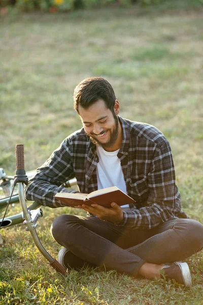 Fröhlicher Typ, der ein Buch liest, auf Gras neben dem Fahrrad im Park sitzt. — Stockfoto