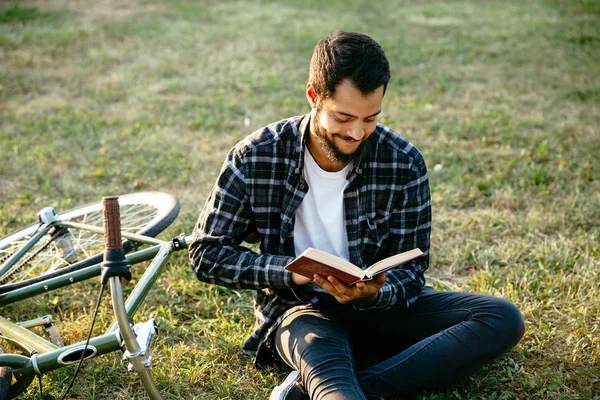 Fröhlicher Mann, der ein Buch liest, neben dem Fahrrad sitzt, im Freien. — Stockfoto