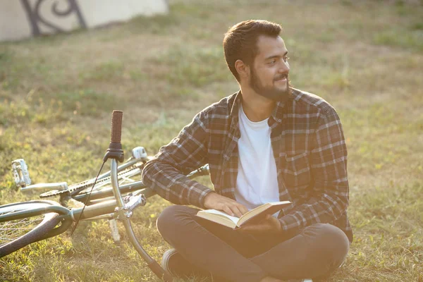 Barbudo leyendo un libro, sentado en la hierba cerca de la bicicleta en el parque . — Foto de Stock