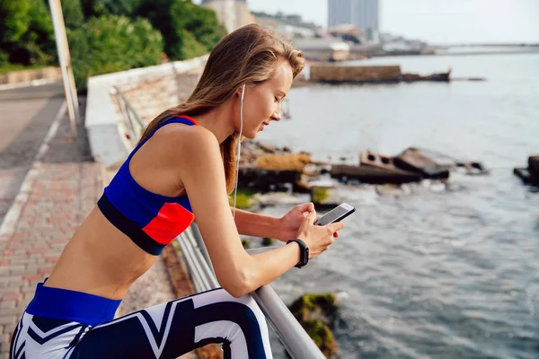 Deportiva con auriculares usando un smartphone, parada en el muelle — Foto de Stock