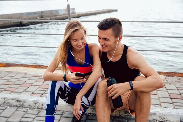 Atleta pareja viendo un video en el teléfono celular, mientras está sentado en el suelo, cerca del mar — Foto de Stock