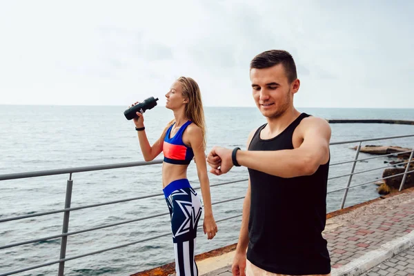 Deportista sonriente revisando un momento después del entrenamiento mientras su novia bebe un agua al aire libre — Foto de Stock