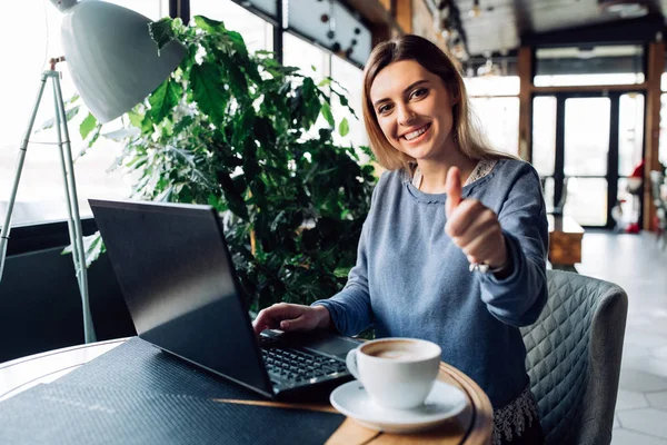 Hermosa chica mostrando un pulgar hacia arriba sentado en la cafetería con el ordenador portátil — Foto de Stock