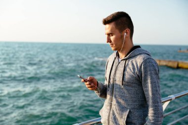 Kulaklıklı sportif adam akıllı telefon kullanarak, deniz kenarında yürüyor