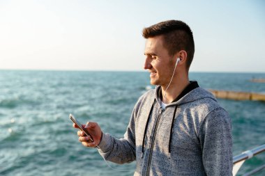 Cep telefonu kullanarak kulaklıkla gülümseyen sporcu, deniz kenarında yürüyor